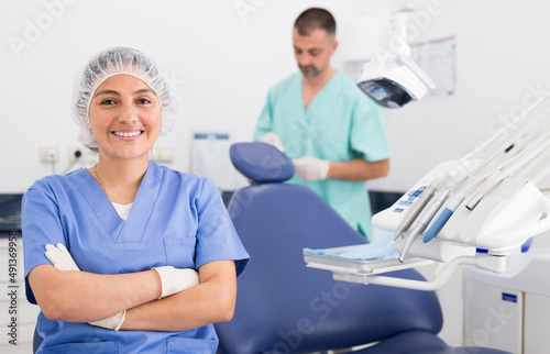 Cheerful latin female dentist in uniform with crossed hands sitting near medical chair