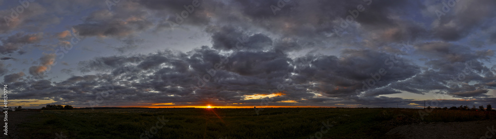 Wide panorama of flat landscape with dark clouds at sunset and orange sun Wide panorama of flat landscape with dark clouds at sunset and orange sun