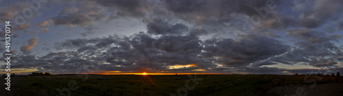Wide panorama of flat landscape with dark clouds at sunset and orange sun