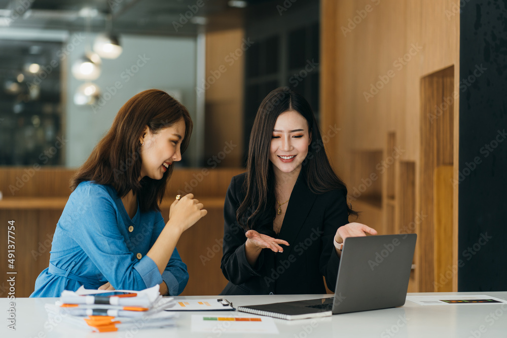Two young women working together in an office at their small business sitting reading a report or paperwork with pleased smiles Two young women working together in an office at their small business sitting reading a report or paperwork with pleased smiles