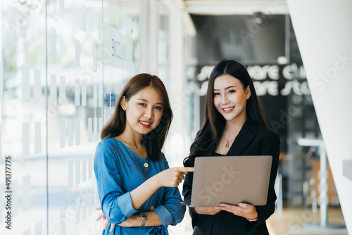 Smiling businesswoman holding digital tablet and talking with partner while standing in modern office interior, team of professional employees discussing ideas of project after working on touch pad