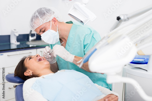 The dentist in face mask inspects female patient teeth with mirror and probe