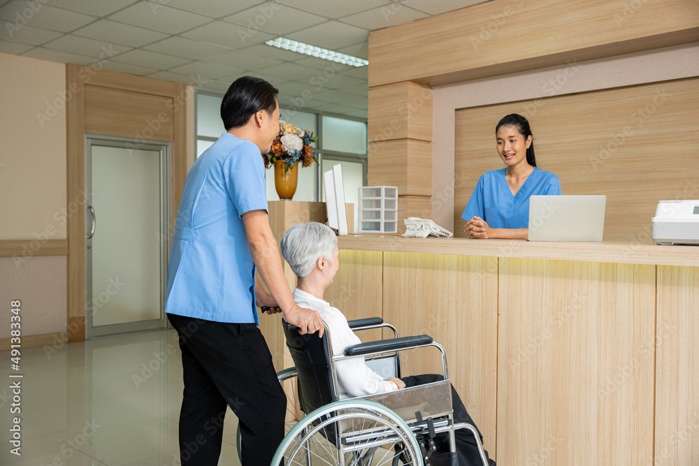 Fototapeta premium Patient elderly sit on wheelchair meet and talking with nurse or staff at front counter in of the hospital, healthcare reception service treatment process concept