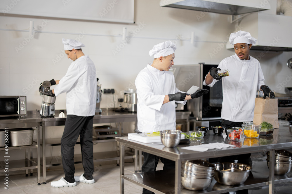 Multiracial team of cooks actively work in the kitchen. Asian chef announces order list from the printed check, Latin guy packs food for deliver, European cooks cooking behind. Concept of teamwork at