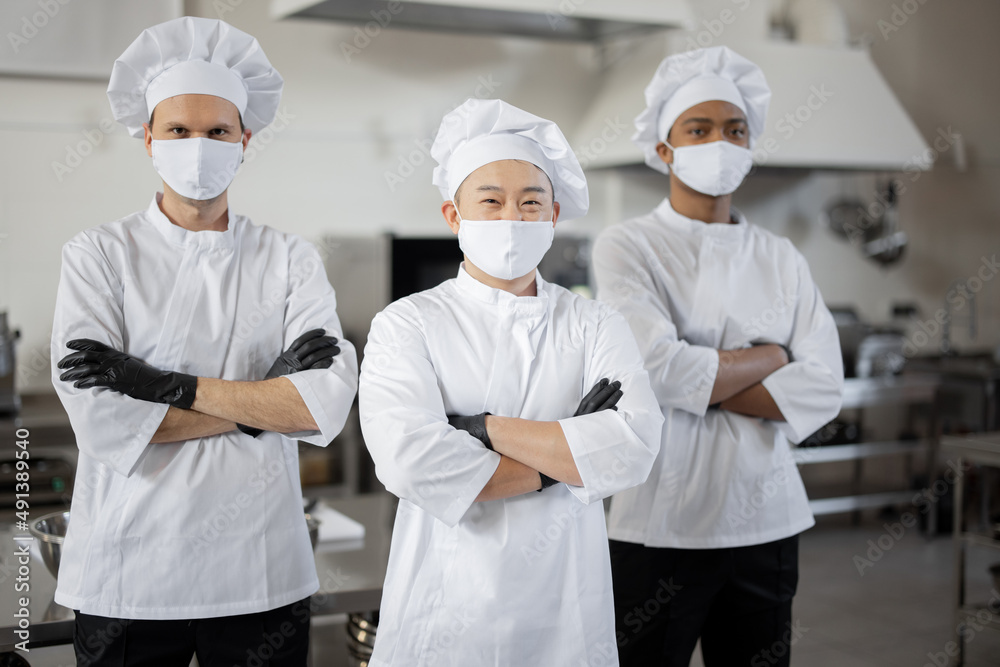 Portrait of multiracial team of three chefs standing together in the professional kitchen. Well-dressed chefs in face masks and protective gloves ready for a job. New normal for business during