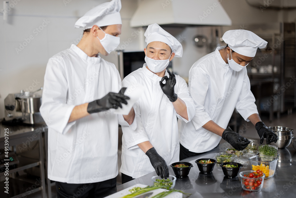 Three well dressed chefs in face masks prepare takeaway food in professional kitchen. Chef reads printed check with the order. Concept of a dark kitchen for cooking for delivery during pandemic