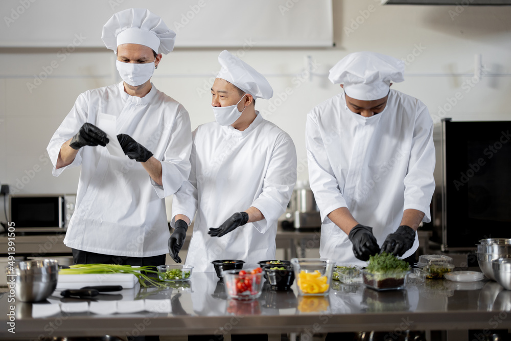 Three well dressed chefs in face masks prepare takeaway food in professional kitchen. Chef reads printed check with the order. Concept of a dark kitchen for cooking for delivery during pandemic