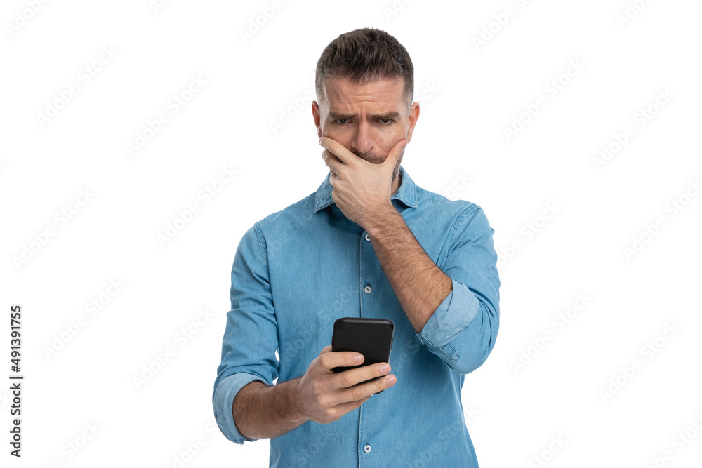 upset young man covering mouth, being disappointed while holding phone upset young man covering mouth, being disappointed while holding phone