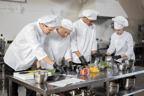 Multiracial team of cooks mixing ingredients for take away food in professional kitchen. Concept of dark kitchen for cooking for delivery. Idea of teamwork in a restaurant. Diverse people at job