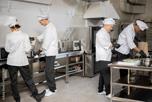 Happy multiracial team of cooks actively work in the kitchen. Asian chef announces order list from the printed check, Latin guy packs food for deliver, European cooks cooking behind. Concept of