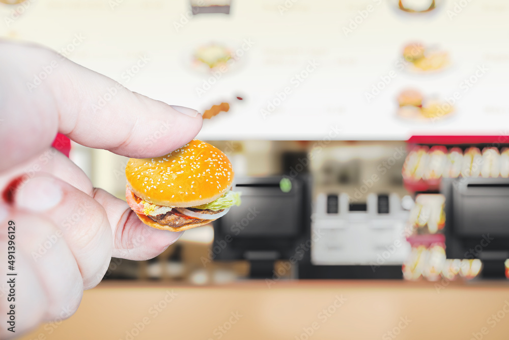 Miniature hamburger in the hand on the background of the diner. Background on the theme of reducing portions in a restaurant and diet.