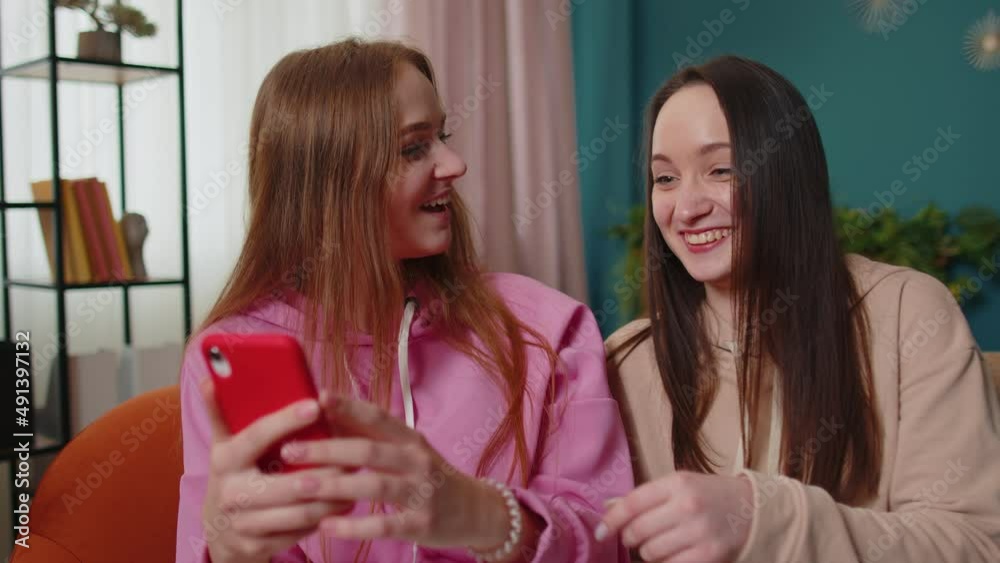 Cheerful girls friends siblings watching funny movies on smartphone, online comedy film. Happy two female women couple family on sofa at home, laughing, spend leisure time on social media network