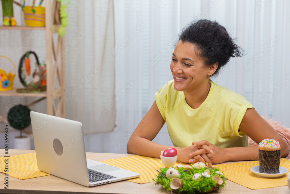 Cute African American woman talking by video call on a laptop. A young woman demonstrates a painted egg and Easter cake while sitting at a decorated holiday table. Happy easter. Close up.