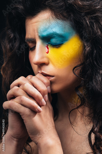 A Ukrainian girl with a bloody tear prays for peace in Ukraine. Make-up in the form of the ensign of Ukraine. War in Ukraine.