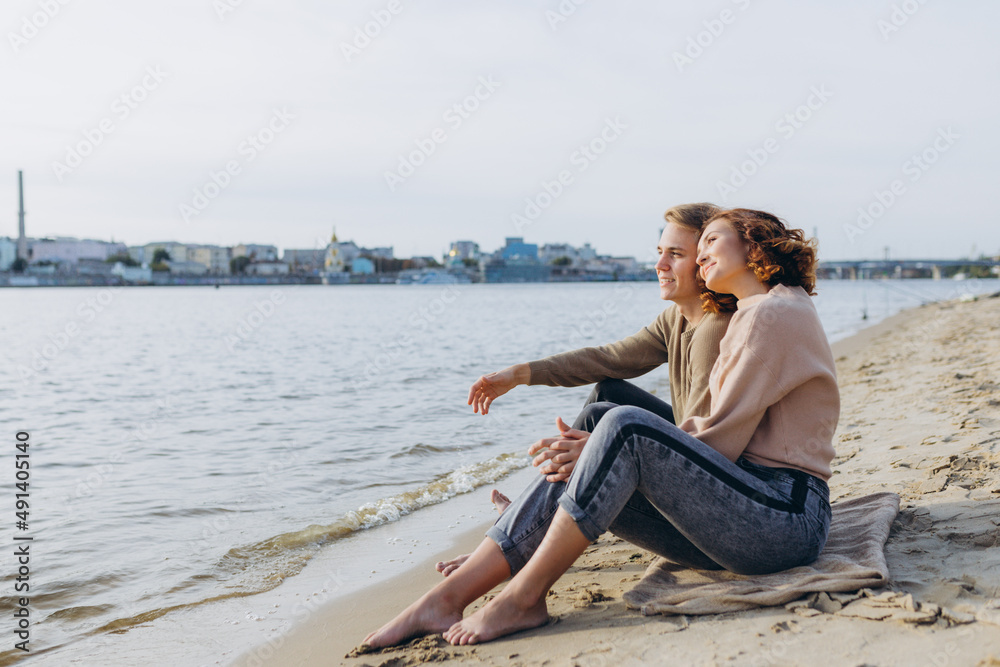 A loving couple has fun - they laugh, hug each other and enjoy a warm summer evening. Romantic couple sitting by the sea. The young guy tells interesting stories of the girl and points aside hands