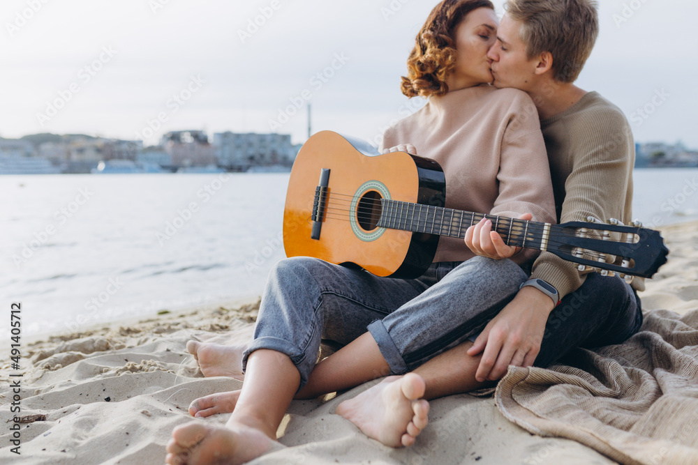 young guy teaching his girlfriend how to play guitar, self-taught. Couple in love having fun on the beach hugging and kissing