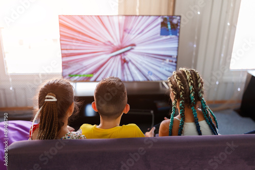 little children a boy and two girls with pigtails play a game console in front of a big TV in the playroom with their backs turned