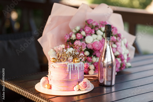 beautiful layered pink cake with golden numbers on a wooden stand twenty five years birthday for a young girl near a large bouquet of roses and a bottle of champagne in a shiny bottle