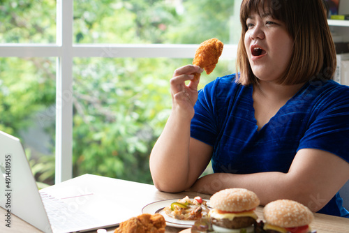 Hungry overweight woman holding Fried Chicken, hamburger on a wooden plate and Pizza on table, During work from home, gain weight problem. Concept of binge eating disorder (BED). photo