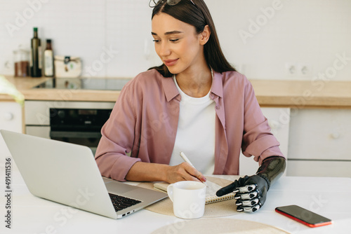 Young cutie with bionic prosthesis instead of hand and glasses on forehead doing homework using laptop, writing in copybook, sitting at table in kitchen in front of smartphone and cup of hot drink photo