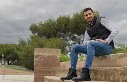 Full length shot of a hispanic cool man sitting on a steps of a viewpoint and looking at the camera while smiling in cloudy day.