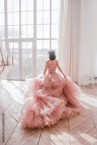 young girl with short black hair in pink princess dress with train is standing from the back near huge panoramic white window background at sunny day. fashion concept, free space