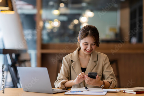 Photo of smiling asian woman using cellphone while working with laptop in office