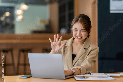 Joyful young woman sit at the work desk, waves hand, greeting colleagues. Attractive business female communicate with colleagues or friends by video call, smiling, remotely work or study © crizzystudio