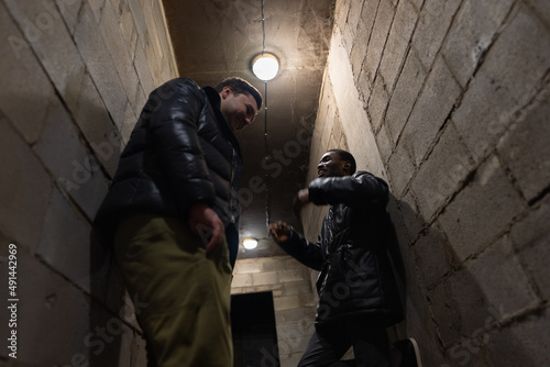 Obraz na płótnie Caucasian guy and black guy are hiding in a bomb-shelter and trying to have fun during russian invasion w salonie