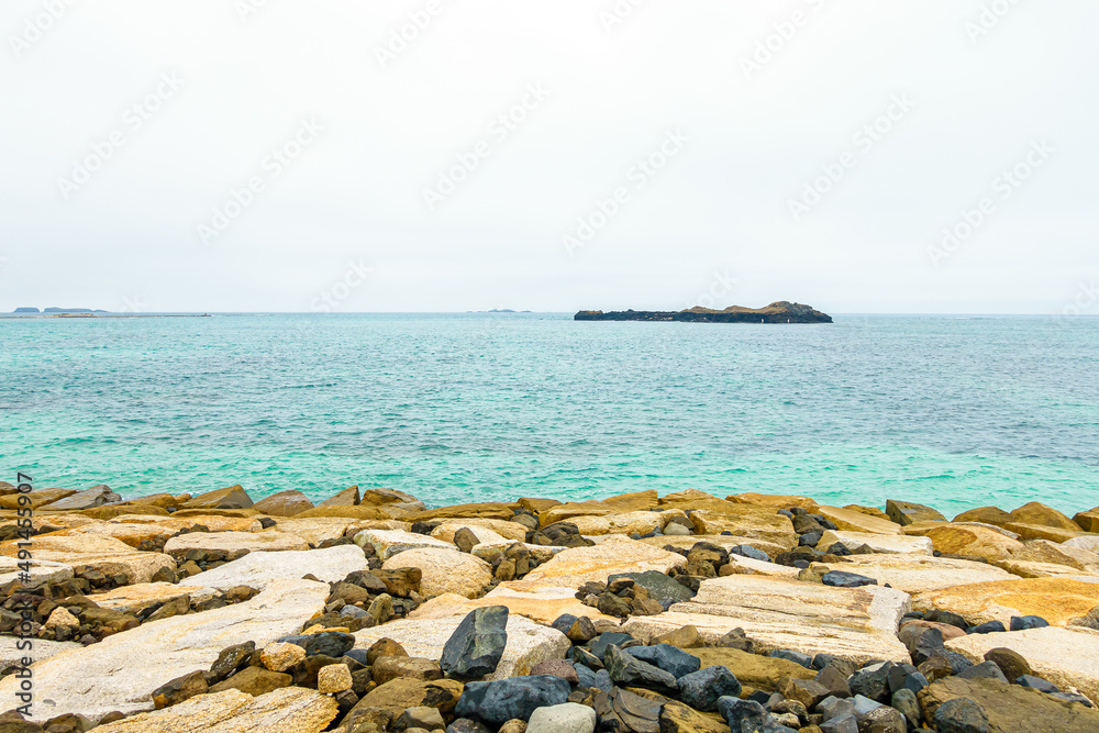 A cloudy day, a coast of stacked yellow rock blocks and a blue gradient ocean, with a small island off the coast