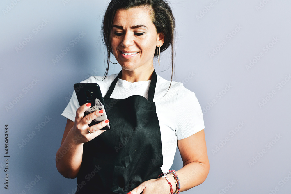 Beautiful Friendly cute Female Barista in black apron Smiling at camera, using mobile Phone to manage cafe orders.Grocery Store employees, small business and Coffee Shops concept. Beautiful Friendly cute Female Barista in black apron Smiling at camera, using mobile Phone to manage cafe orders.Grocery Store employees, small business and Coffee Shops concept.