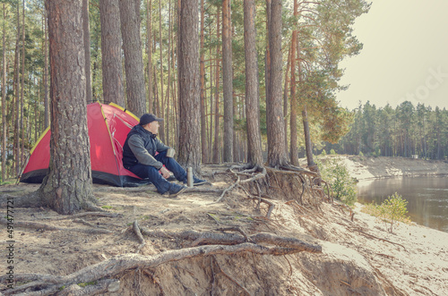 Young man sits near a tent in the forest on a high bank of the river.