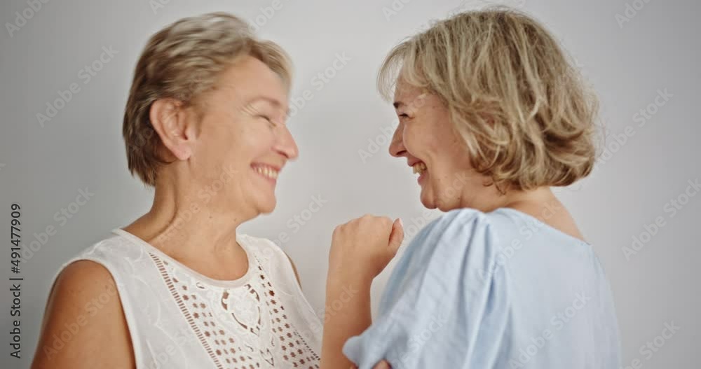 Mother and daughter gossiping and laughing. Blond adult daughter whispering secret to ear of mother then laughing cheerfully against gray background Mother and daughter gossiping and laughing. Blond adult daughter whispering secret to ear of mother then laughing cheerfully against gray background