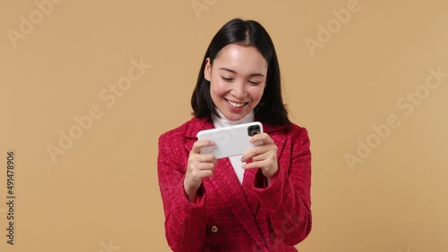 Gambling young woman of Asian ethnicity 20s wears red jacket using play racing on mobile cell phone hold gadget smartphone for pc video games isolated on plain pastel beige background studio portrait photo