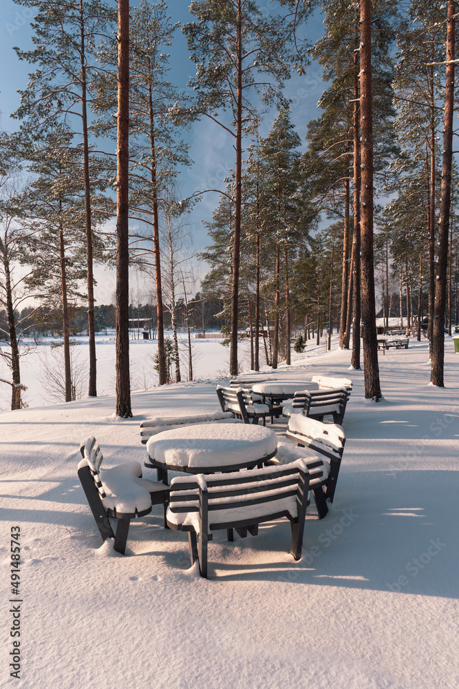 Picnic table by the lake under snow in winter. A thick layer of snow after a snowfall covered benches and tables. Pine forest on a sunny winter day. Latvia
