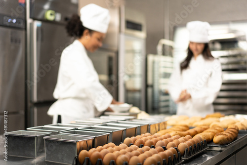 Selective focus of many empty baking bread loaf pans placed near two trays of 30 chicken eggs and a tray of baked breads on a table where blurred two female bakers are kneading dough in a kitchen.