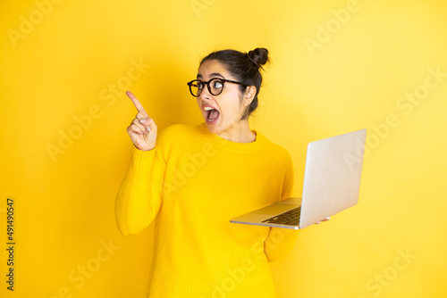 Young brunette woman working using computer laptop over yellow background amazed and pointing with hand and finger to the side