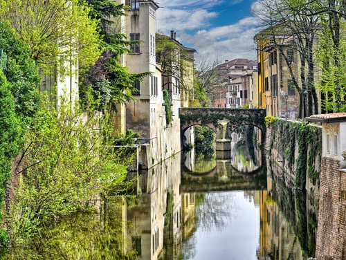  En Modena (Italia) un angosto rio o canal atravesando dos barrios unidos por un bello puente en desnivel que se refleja en el agua.