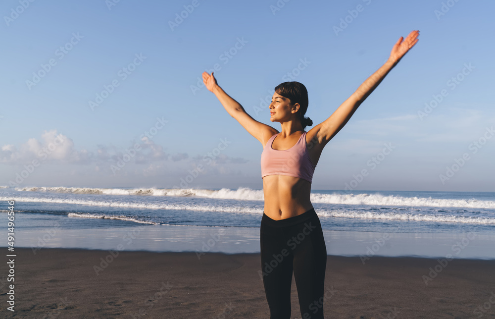 Attractive female yogi in black leggins and pink top raising hands looking at sunshine spending morning at coastline, Caucasian woman in sportswear warm up for feeling vitality and wellness Attractive female yogi in black leggins and pink top raising hands looking at sunshine spending morning at coastline, Caucasian woman in sportswear warm up for feeling vitality and wellness