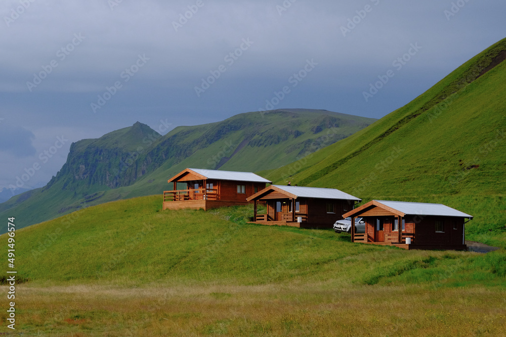 3 wooden country houses (cottage village) stand next to the mountain on the background of mountains covered with green grass in Iceland. Landscape with tourist houses (hotels) in Iceland
