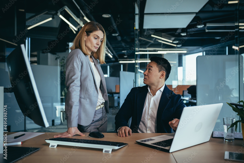 happy coworkers discuss project strategy by looking and pointing at laptop pc computer monitor screen. multiethnic business team in the office. confident mature asian man explaining young female happy coworkers discuss project strategy by looking and pointing at laptop pc computer monitor screen. multiethnic business team in the office. confident mature asian man explaining young female
