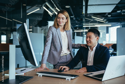 Female boss flirts with male employee, businesswoman sitting at desk and smiling then Asian