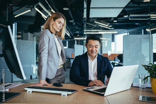 happy coworkers discuss project strategy by looking and pointing at laptop pc computer monitor screen. multiethnic business team in the office. confident mature asian man explaining young female