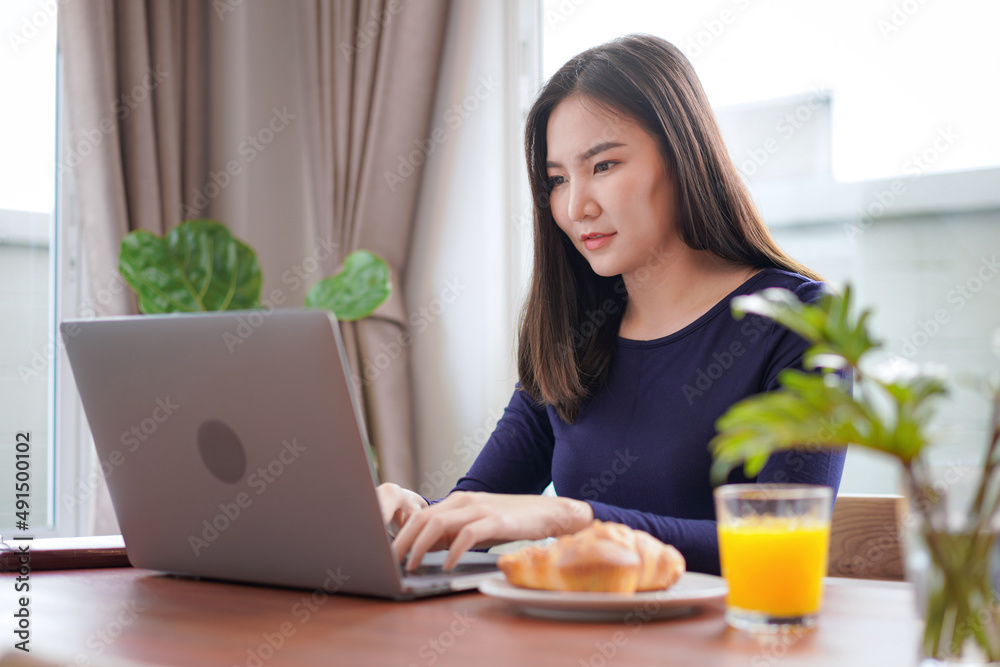 Young asian woman eating bread with a glass of orange juice while looking on laptop screen to typing