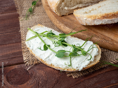 Close-up of a still life made of fresh crusty bread. A slice of bread smeared with cottage cheese and decorated with micro greenery. Linen napkin brown wooden background, top view, flat lay