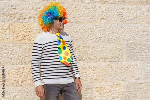 Man dressed up as a Halloween or Purim clown with a rainbow wig and red nose. Portrait of senior male with colorful wig and tie. Funny old man in a clown costume at Purim Jewish Holiday celebration.