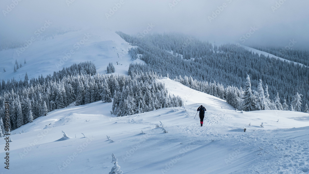 Portrait of a young hiker in the distance dressed all in black climbing the uphill full of snow. Winter season and the sun shining in the background