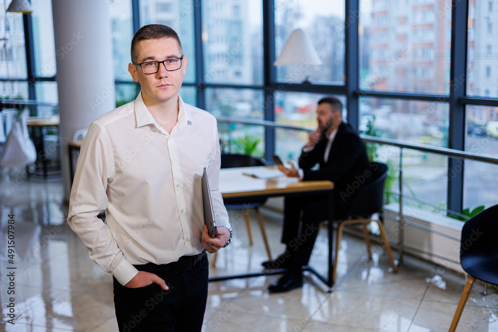 A young businessman man in glasses and a white shirt with a laptop in his hands stands in the office and works A young businessman man in glasses and a white shirt with a laptop in his hands stands in the office and works