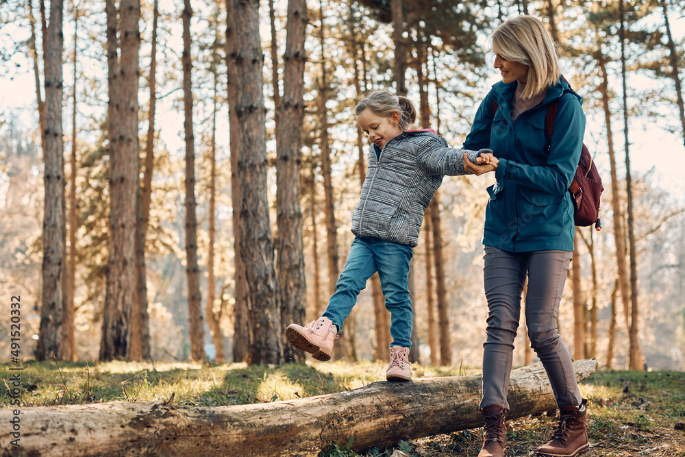 Happy mother assisting her daughter in maintaining balance while walking on tree trunk in nature.