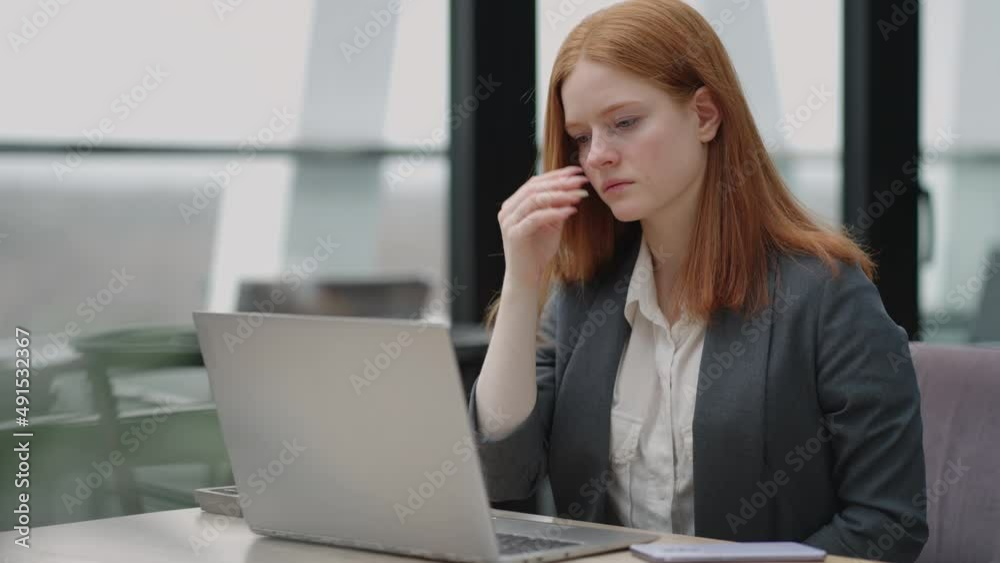 A young red-haired business woman looks thoughtfully at the screen and brainstorms. Watch and think about problems looking out the window. Thoughtful business woman A young red-haired business woman looks thoughtfully at the screen and brainstorms. Watch and think about problems looking out the window. Thoughtful business woman